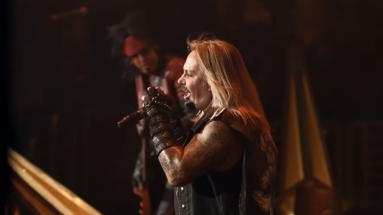 A blond, tattooed rock musician performs with a microphone onstage