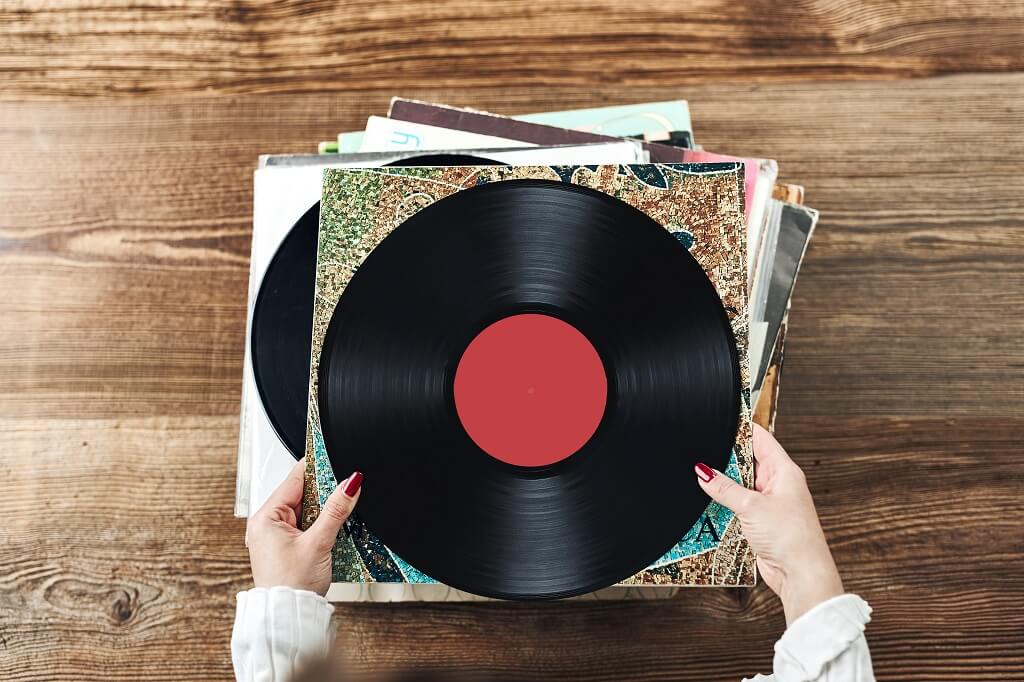 Hands holding vinyl record above a stack of albums on a wooden surface
