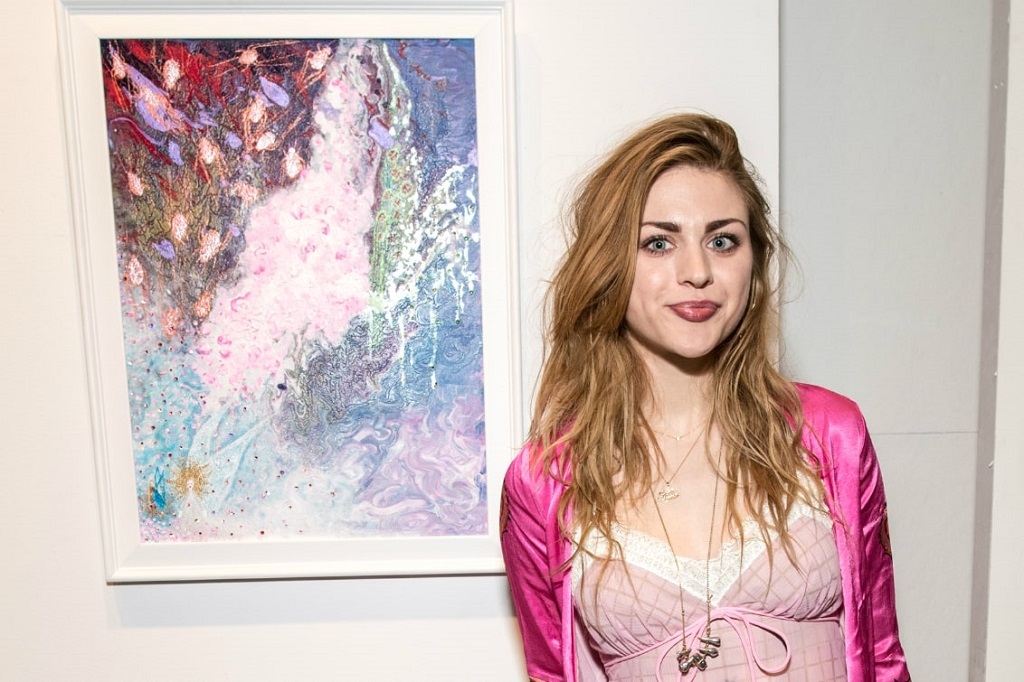 Kurt Cobain’s daughter standing beside her abstract painting at an art gallery, wearing a pink outfit and smiling