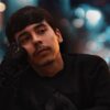 Young man in a black outfit resting his face on his hand with blurred city lights in the background
