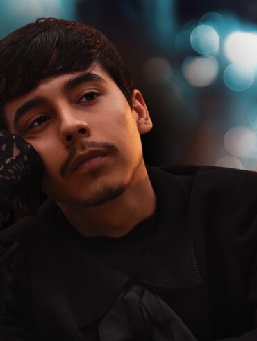 Young man in a black outfit resting his face on his hand with blurred city lights in the background