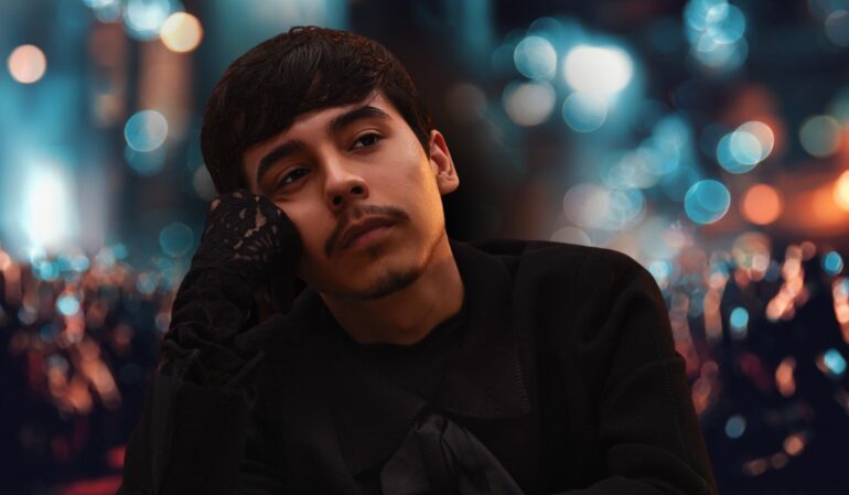 Young man in a black outfit resting his face on his hand with blurred city lights in the background