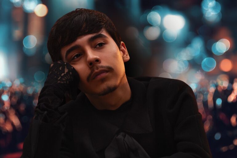 Young man in a black outfit resting his face on his hand with blurred city lights in the background
