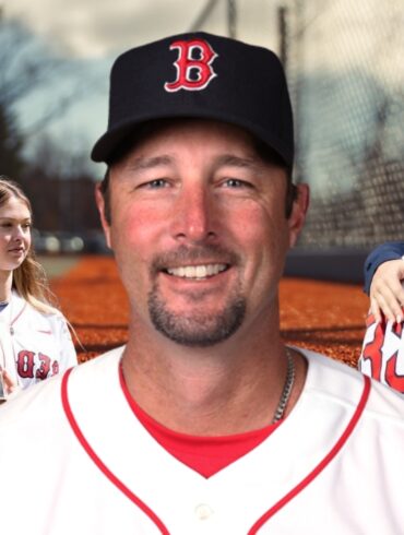 Baseball player in a Red Sox uniform smiling, with family and a championship trophy in the background