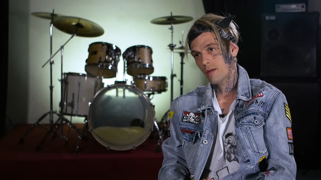 Aaron Carter in a denim jacket with patches, sitting in front of a drum set, with face and neck tattoos visible