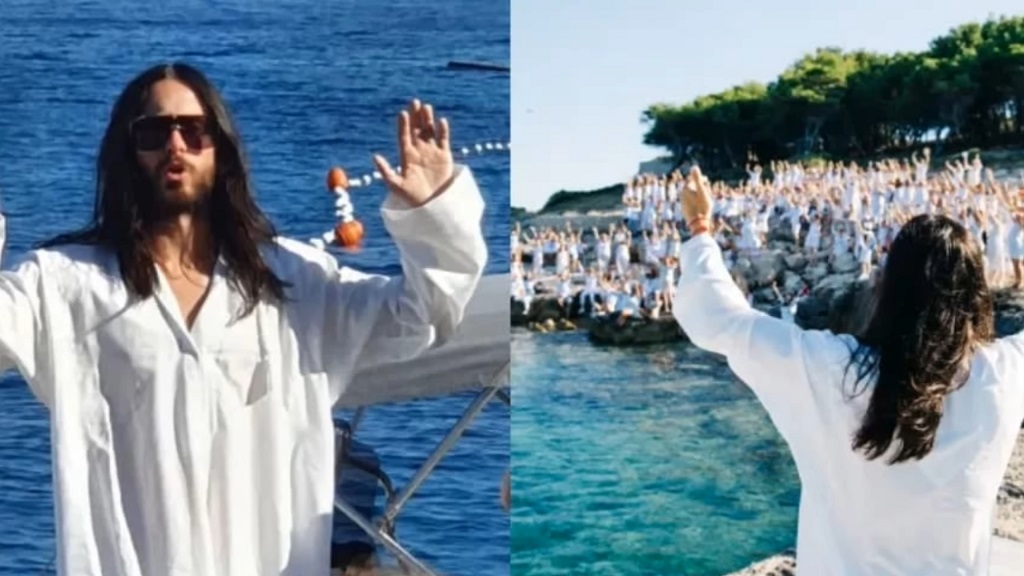 Jared in a white robe raising his hands by the sea, with a large group of people in white gathered on the rocky shore behind him.