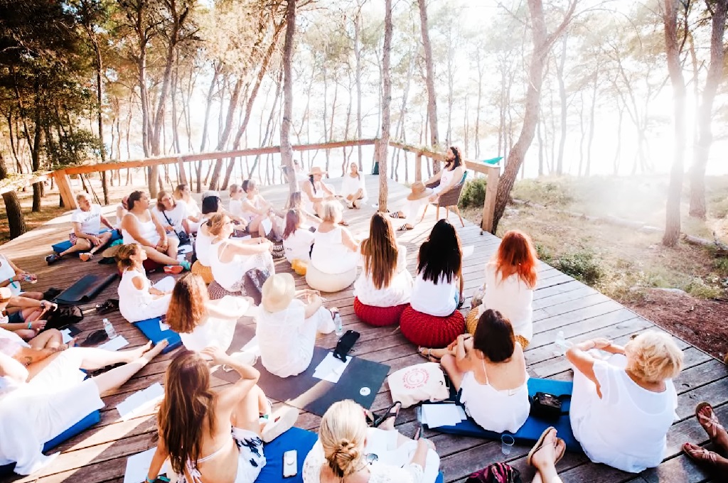 Group of people dressed mostly in white sitting on mats in a wooded outdoor setting, listening to a speaker at the front