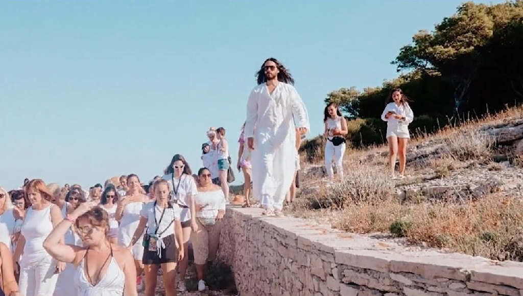 Man in flowing white clothes walking along a stone wall, surrounded by a group of festivalgoers dressed mostly in white, near the Adriatic coast