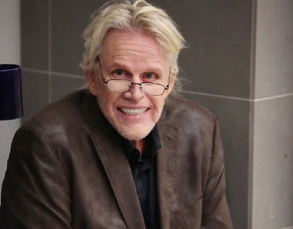 Gary Busey wearing glasses and a brown leather jacket, smiling while seated indoors