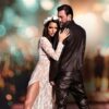 Man in black leather outfit with a woman in a sheer white lace dress and floral crown posing against a blurred background.