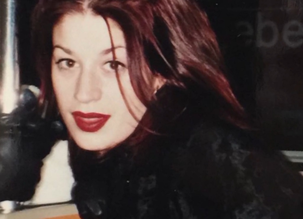 Jennifer Syme with red lipstick and auburn hair, wearing a black outfit and gloves, looking directly at the camera