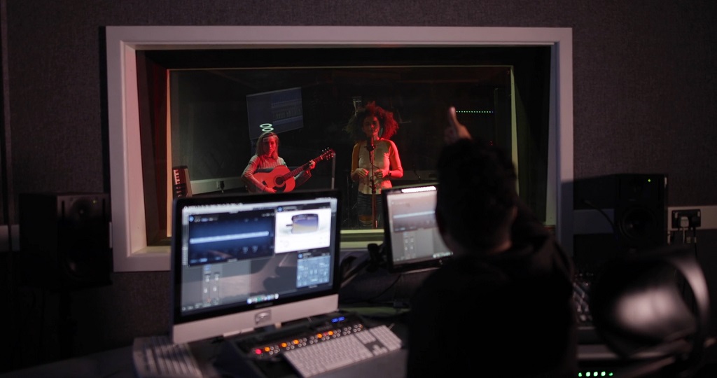 Recording studio with a sound engineer at a mixing desk while two musicians, one singing and one playing guitar, perform in the booth