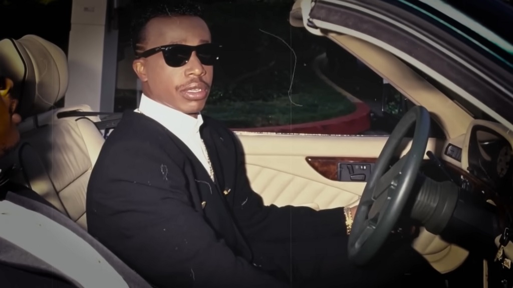 MC Hammer wearing sunglasses and a suit while sitting in the driver’s seat of a luxury car