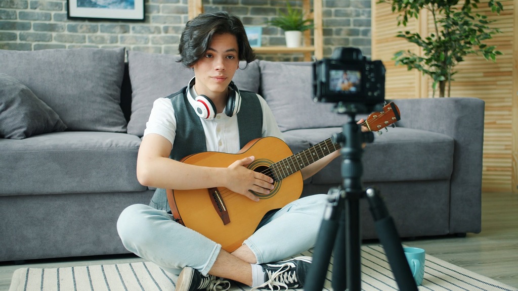 Young musician with headphones around his neck playing guitar while recording a video at home