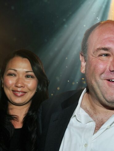 Who Was James Gandolfini’s Wife? Remembering Deborah Lin’s Private Life Actor James Gandolfini smiling in a suit with his wife Deborah Lin standing beside him at an event, illuminated by stage lights