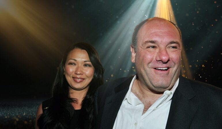 Actor James Gandolfini smiling in a suit with his wife Deborah Lin standing beside him at an event, illuminated by stage lights