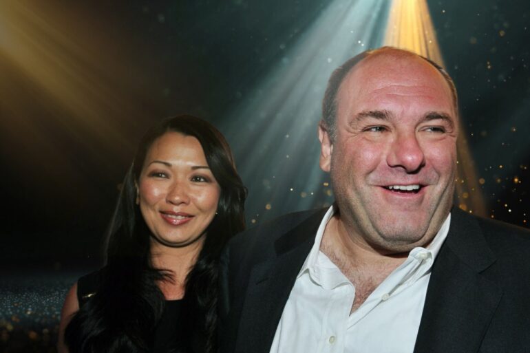 Actor James Gandolfini smiling in a suit with his wife Deborah Lin standing beside him at an event, illuminated by stage lights