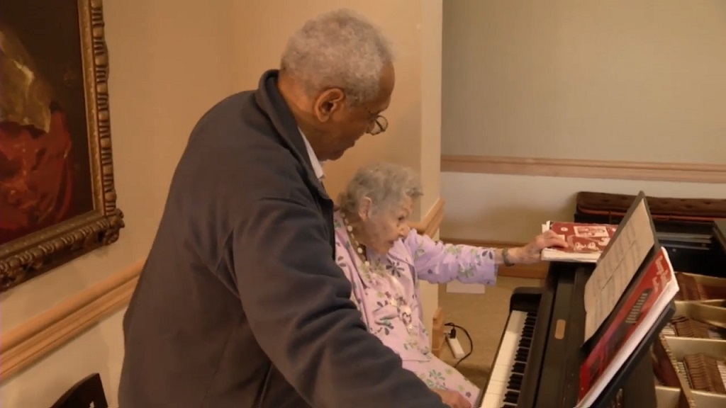 The Final Encore: Retired Celebrities Find a Stage at the Lillian Booth Actors' Home An elderly man helps an elderly woman at the piano during a music activity in a senior living home