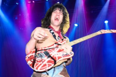 van Halen shirtless on stage under vibrant blue and magenta spotlights, holding a striped red and white electric guitar