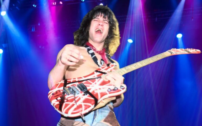 van Halen shirtless on stage under vibrant blue and magenta spotlights, holding a striped red and white electric guitar
