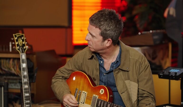 Noel Gallagher sits in a studio holding his guitar during a recording session
