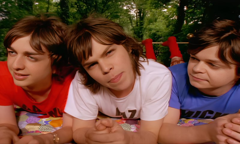 Three young men lie on a colorful blanket outdoors, wearing red, white, and blue shirts