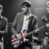 Three musicians perform on stage in suits. The singer holds a vintage mic, the guitarist plays a UK flag guitar