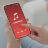 A person holding a smartphone with a red screen displaying a music note icon and the word "STREAMING."