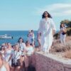 A person in a flowing white robe leads a group, all dressed in white, along a stone path by the sea