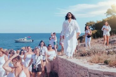A person in a flowing white robe leads a group, all dressed in white, along a stone path by the sea