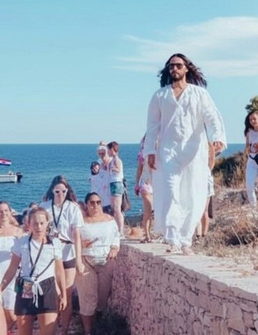 A person in a flowing white robe leads a group, all dressed in white, along a stone path by the sea