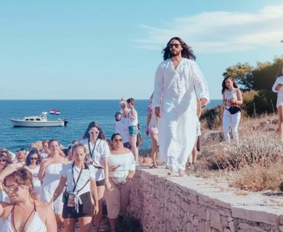 A person in a flowing white robe leads a group, all dressed in white, along a stone path by the sea