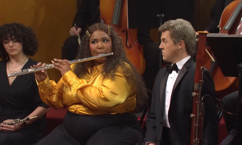 How Lizzo Helped Make Classical Training Feel Cool Again Lizzo in a bright yellow blouse plays the flute energetically in an orchestra