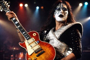 A musician with dramatic black-and-white face paint and a shiny, silver costume plays an electric guitar on stage