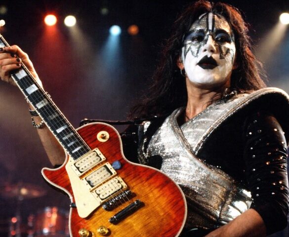 A musician with dramatic black-and-white face paint and a shiny, silver costume plays an electric guitar on stage
