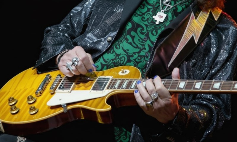 The Life and Career of Ace Frehley, the Guitarist Who Helped Shape the Sound of KISS A musician strums a sunburst electric guitar, wearing rings and a glittering jacket