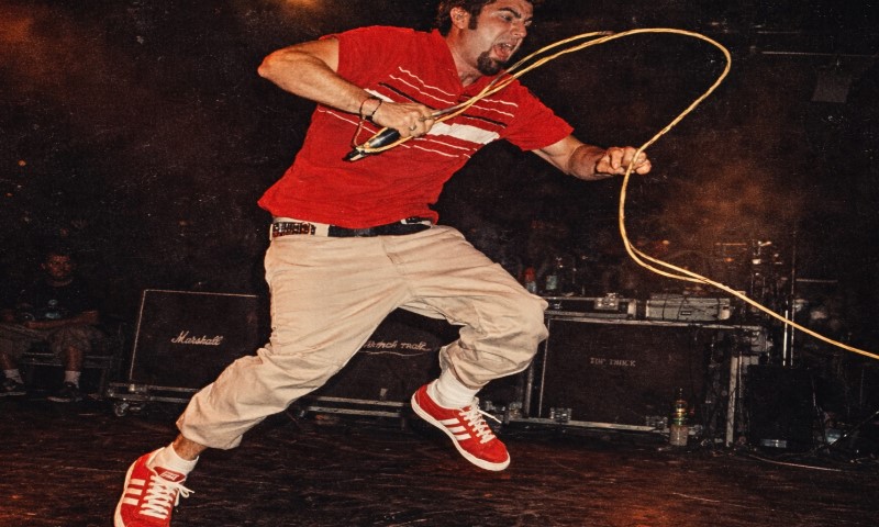 A man mid-jump on stage, wearing a red shirt and beige pants, energetically swinging a microphone