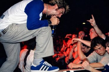 A male performer with spiky hair passionately sings into a microphone at a concert, leaning toward an enthusiastic crowd