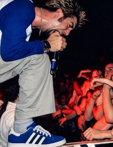 A male performer with spiky hair passionately sings into a microphone at a concert, leaning toward an enthusiastic crowd