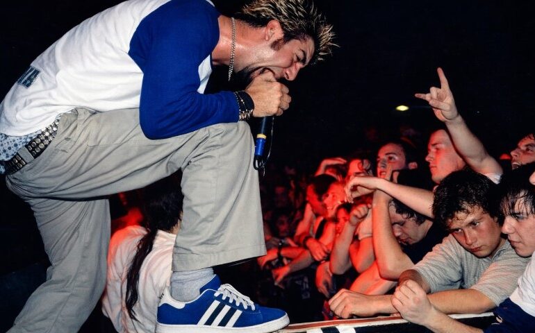 A male performer with spiky hair passionately sings into a microphone at a concert, leaning toward an enthusiastic crowd