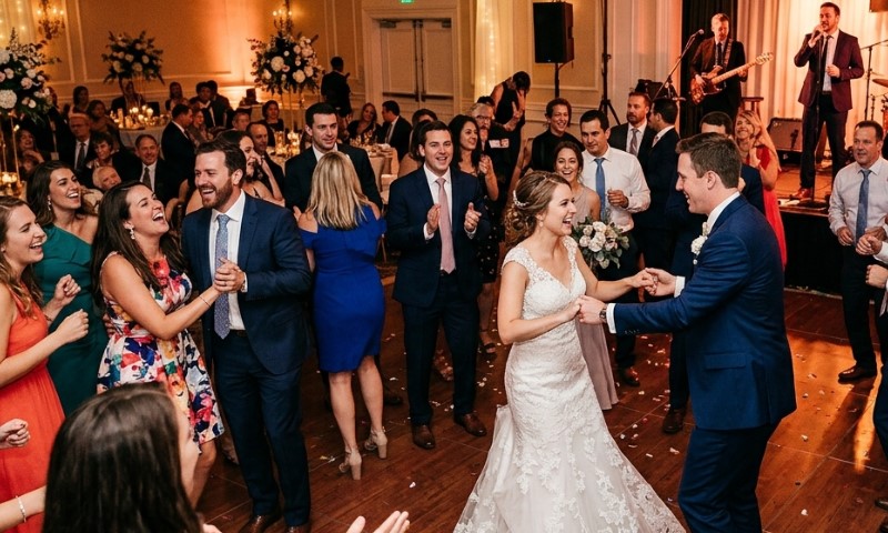 Best Songs For A Modern Wedding: Music Ideas Guests Will Enjoy A bride and groom joyfully dance in the center of a lively reception, surrounded by smiling guests clapping along