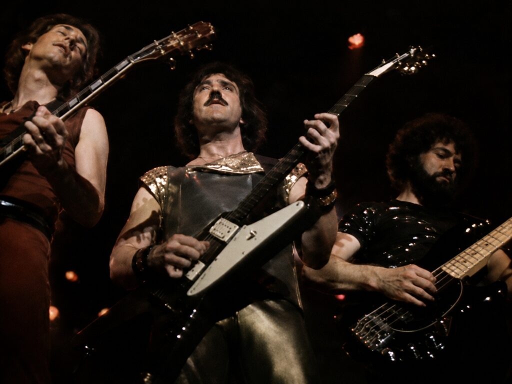 10 Rock and Metal Bands Inspired by Fantasy Books Three rock musicians playing guitars during a live Blue Öyster Cult concert.