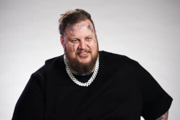 Jelly Roll smiles in a black shirt with a silver chain against a plain gray backdrop