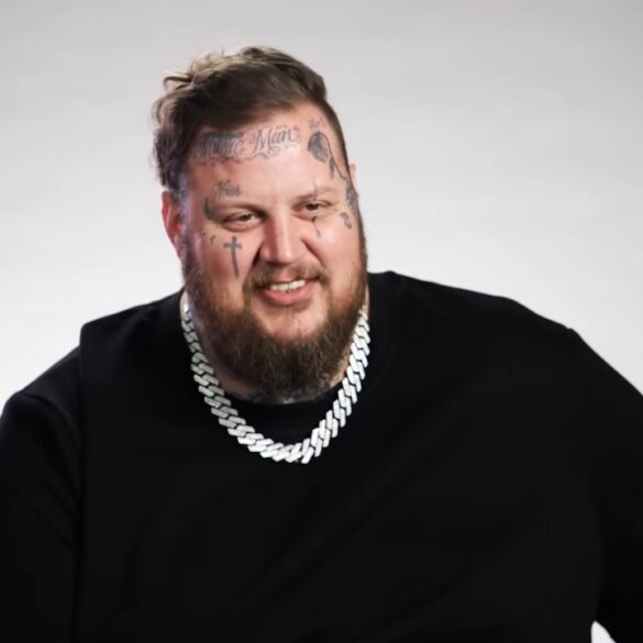 Jelly Roll smiles in a black shirt with a silver chain against a plain gray backdrop