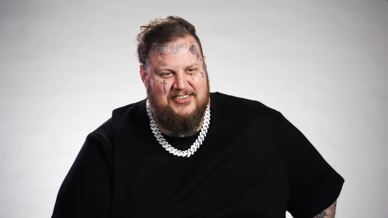 Jelly Roll smiles in a black shirt with a silver chain against a plain gray backdrop