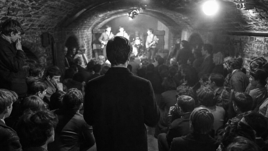 The Biggest Myths About How Famous Rock Bands Were Discovered A man in an overcoat watches a band play to a packed brick cellar club