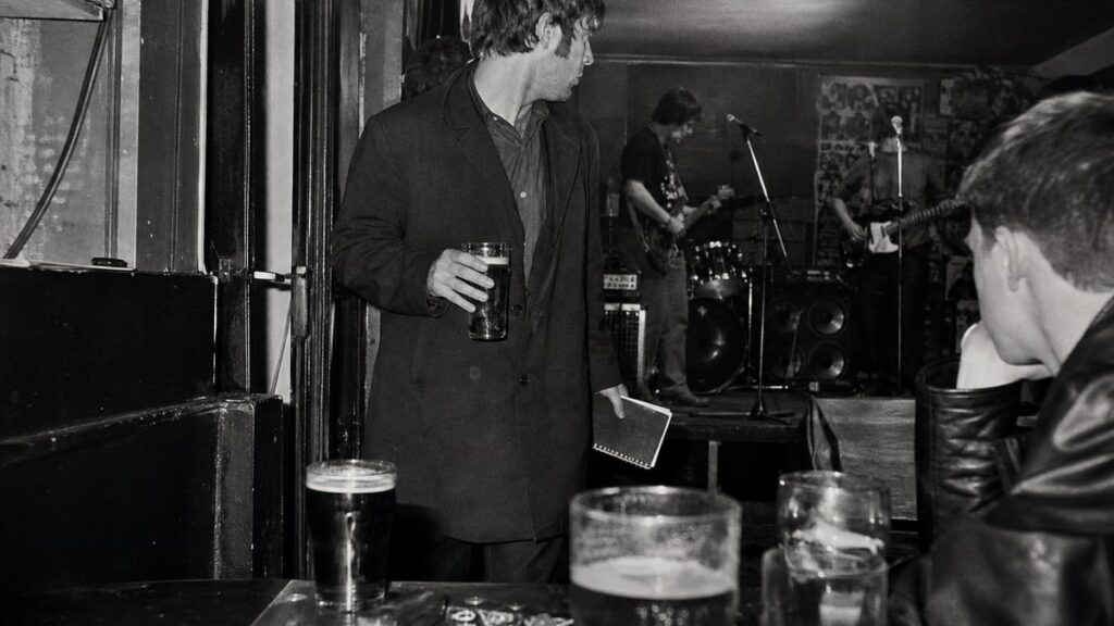 The Biggest Myths About How Famous Rock Bands Were Discovered A record label figure pauses inside a crowded pub while musicians play on a small stage