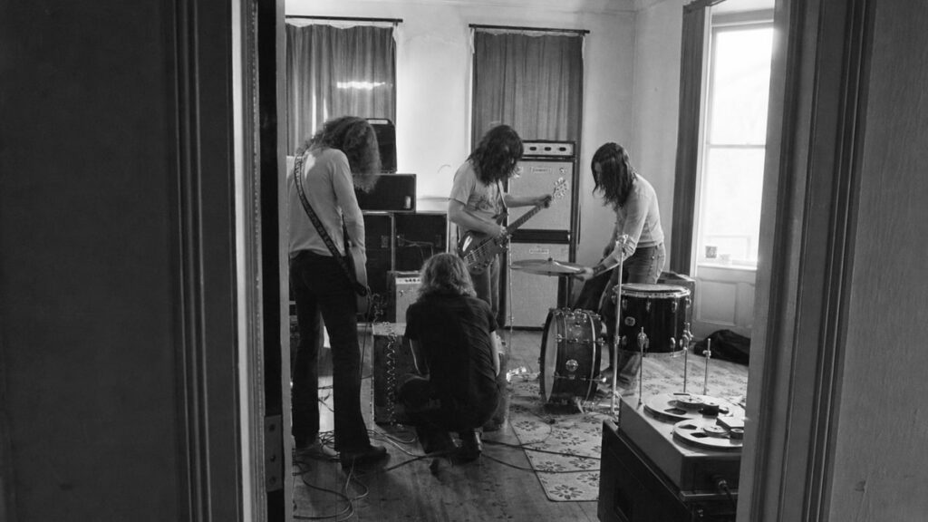 The Biggest Myths About How Famous Rock Bands Were Discovered Four musicians tune instruments in a sparse rehearsal room, seen through an open doorway