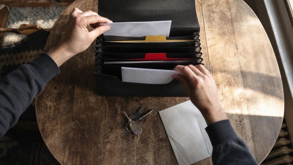 The 10 Essentials to Deal With Right After Buying a House, Design Changes Can Wait Hands sort blank papers into a black file box on a dining table beside house keys.