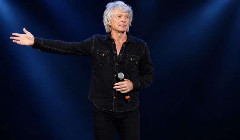 Jon Bon Jovi with silver hair and a black shirt holds a microphone and extends his arm against a dark blue concert background.